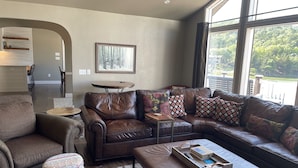 Ein Wohnzimmer mit einem braunen Ledersofa, einem gemusterten Ottomanen und einem Couchtisch mit Tablett. An der Wand hängt ein gerahmtes Bild, und es gibt einen kleinen runden Tisch sowie einen Blick auf eine Terrasse durch große Fenster.