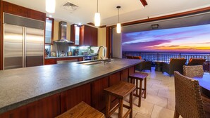 Een moderne keuken met een groot kookeiland, roestvrijstalen apparatuur en uitzicht op de zonsondergang boven de oceaan.