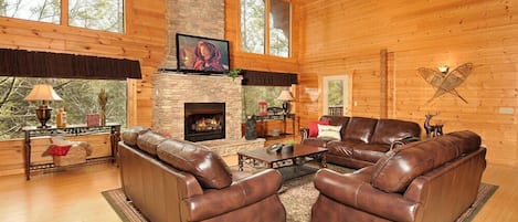 A spacious living room with leather sofas, a fireplace, and large windows offering a view of the outdoors.