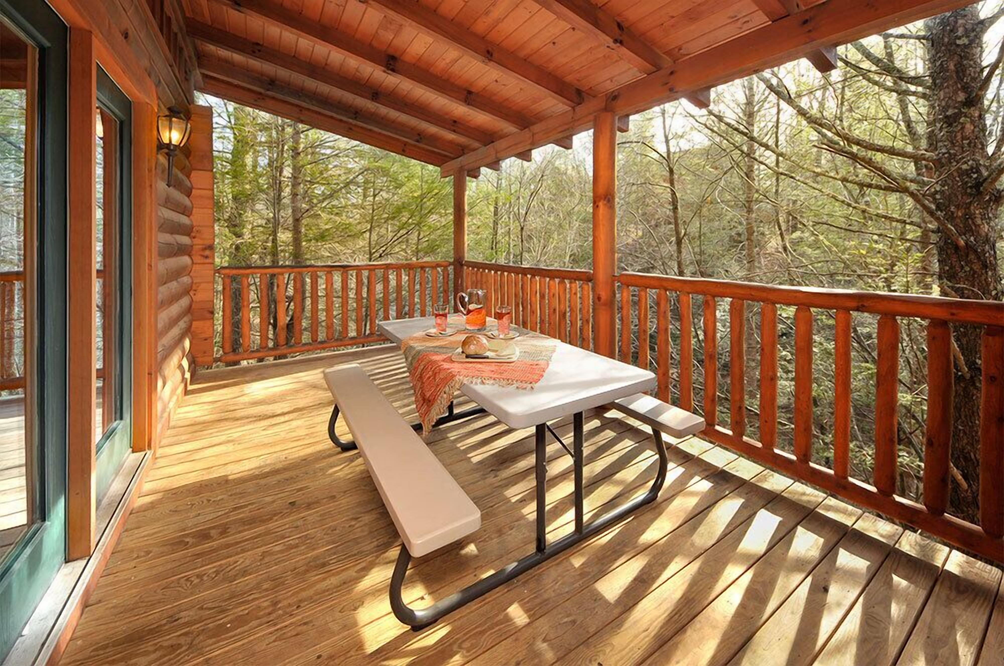 A wooden deck with a picnic table and benches, surrounded by a wooden railing, overlooking a forested area.