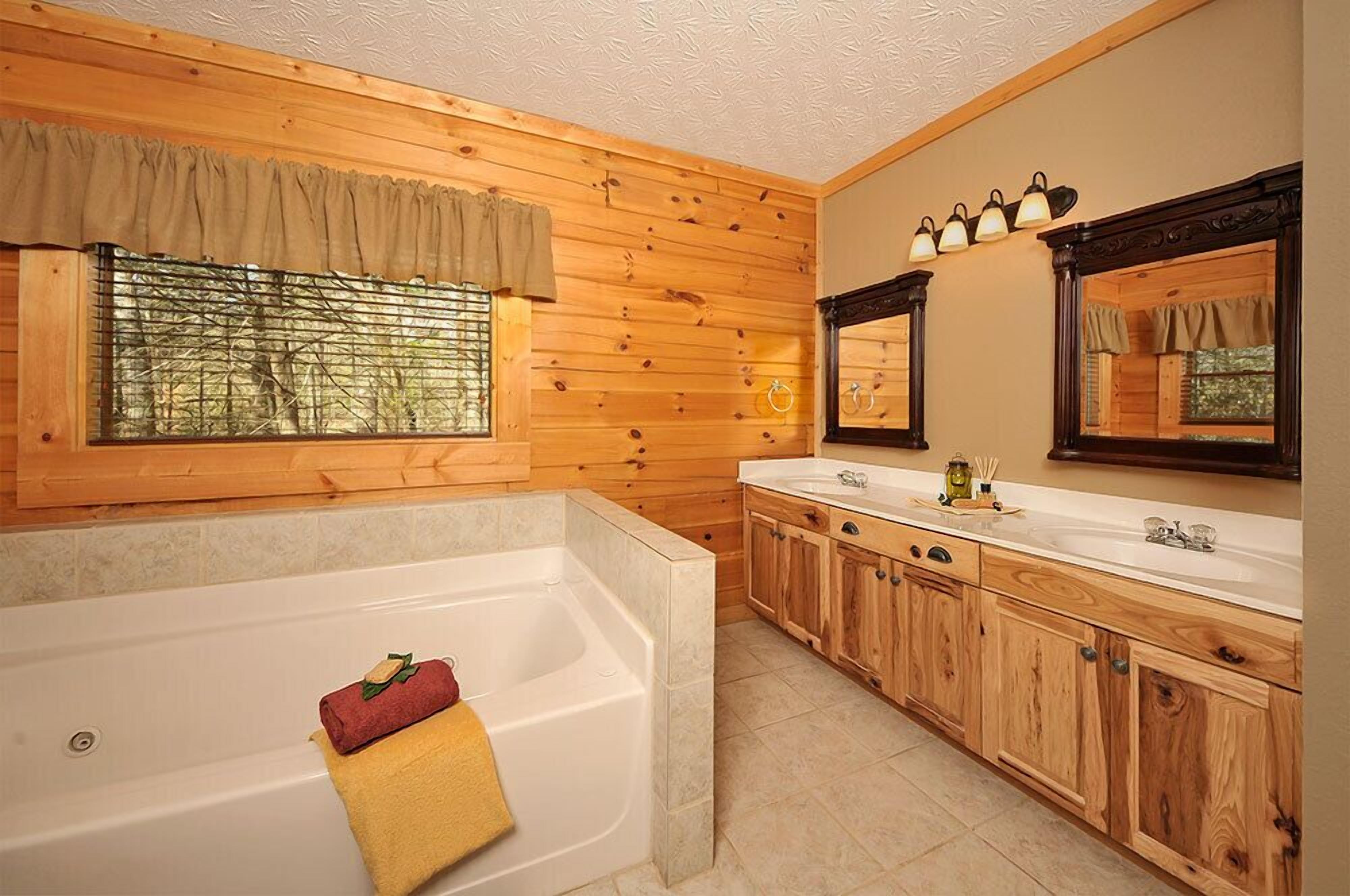 A bathroom with wooden paneling, a large bathtub, a double sink vanity, and a window with a view of trees.