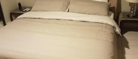 A neatly made bed with a beige comforter and pillows, a bedside table with a lamp, and a telephone.