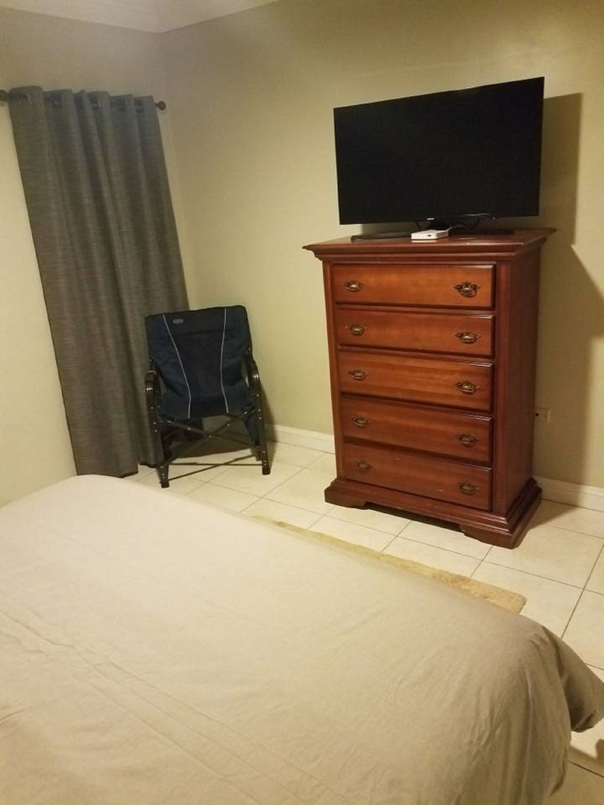 A bedroom with a bed, a wooden dresser with a television on top, a chair, and a curtain.
