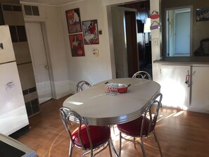 A dining area with a white table, red chairs, and a red Coca-Cola pan. There are wall-mounted pictures and a refrigerator in the background.