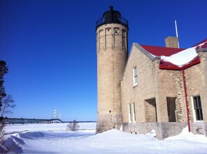 A brick lighthouse with a red roof, a clear blue sky, and a bridge in the background.