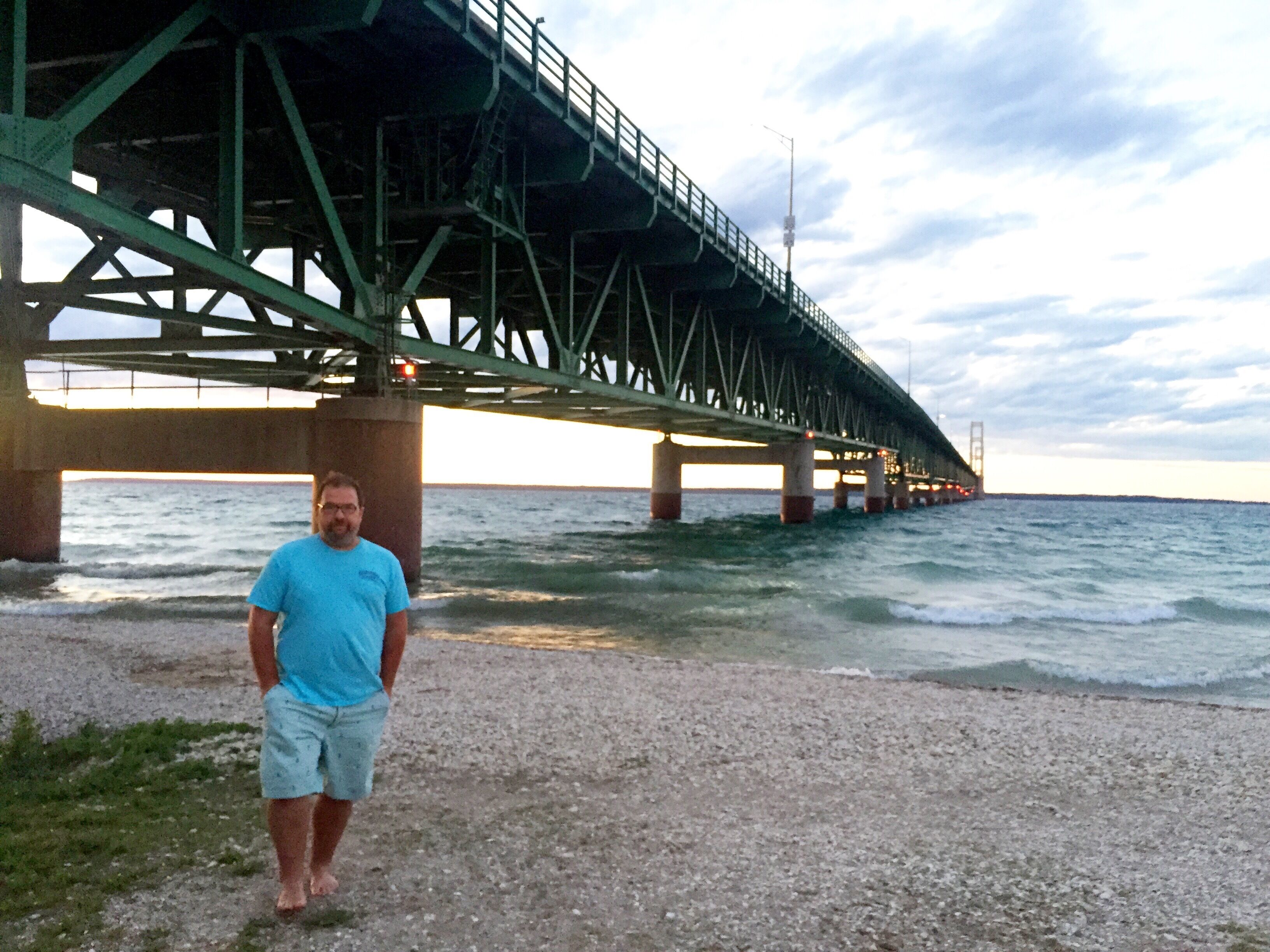 A man standing on a gravel path under a bridge with the ocean in the background.