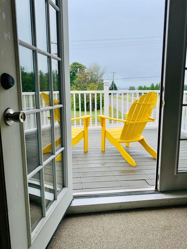A view through a glass door to a deck with a yellow Adirondack chair and a small table.