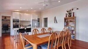 A dining area with a wooden table and chairs, a kitchen in the background, and a bookshelf with books and decorative items. - Esperance Landing, waterfront luxury (Brooks Bay)
