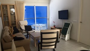 En kompakt opholdsstue med sofa, lille bord, stol, fjernsyn og et vindue med persienner.