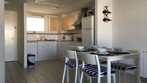 Et lille køkken med hvide skabe, indbygget mikroovn og et køleskab. Der er et spisebord med stole og en skraldespand.