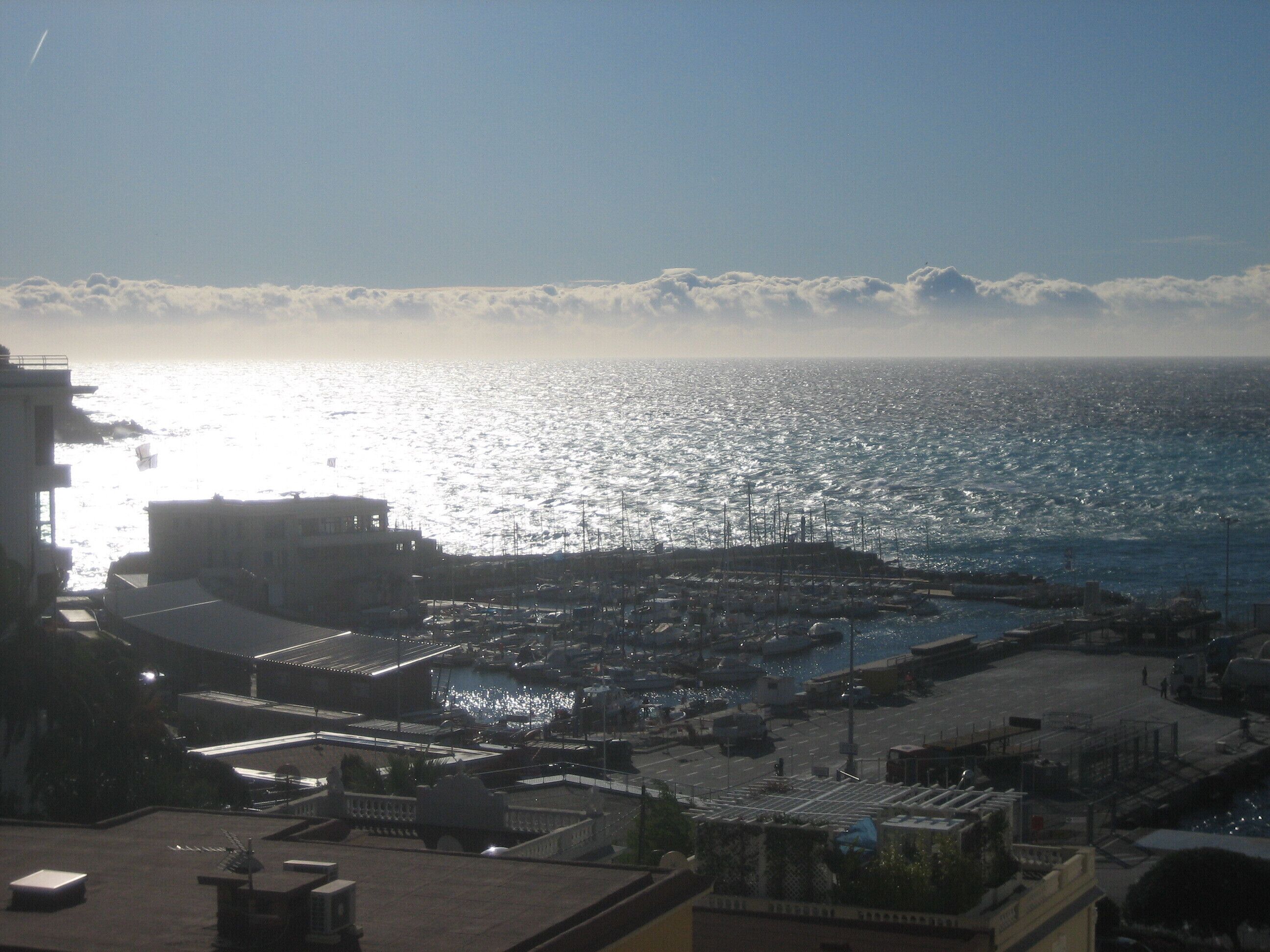Uma marina costeira com barcos atracados, edifícios em primeiro plano e o oceano ao fundo.