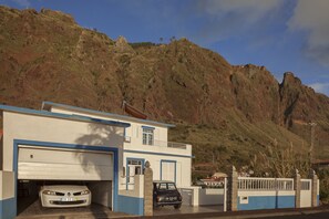 A building with a garage, a car parked inside, and a mountainous backdrop. - Holiday house by the sea. (Calheta/ Madeira)