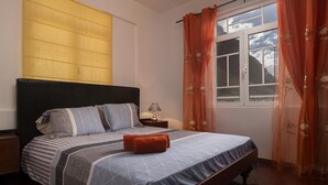 A neatly made bed with a geometric patterned bedspread, a dark headboard, and a window with orange curtains. - Holiday house by the sea. (Calheta/ Madeira)