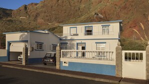 A two-story building with a garage, a balcony, and a mountain backdrop. - Holiday house by the sea. (Calheta/ Madeira)