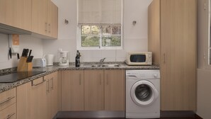 A modern kitchen with wooden cabinets, a built-in microwave, a washing machine, and a window with blinds. - Holiday house by the sea. (Calheta/ Madeira)