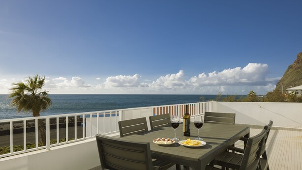 A dining table with chairs and plates of food, a bottle of wine, and a glass of wine on a balcony overlooking the ocean. - Holiday house by the sea. (Calheta/ Madeira)