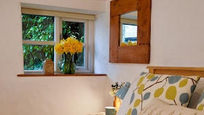 Un dormitorio con cama, mesita de noche, ventana con vistas a la vegetación, un florero con flores y un espejo.