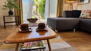 Una sala de estar acogedora con una mesa de centro de madera, dos tazas, un bol y una revista. Hay un sofá gris con un cojín estampado, una planta en maceta y una lámpara sobre una mesita.