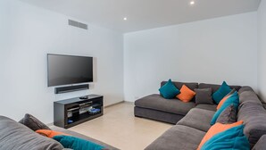 A modern living room with a flat-screen TV mounted on the wall, a grey sofa with teal and orange cushions, and a black entertainment unit with a Blu-ray player.