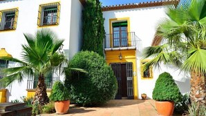 Ein weißes Gebäude mit gelben Verzierungen und einem Balkon. Im Vordergrund stehen Kübelpflanzen und eine Palme.
