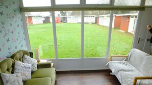 Une pièce avec un canapé vert, un canapé blanc et une chaise en bois. Une grande fenêtre offre une vue sur une cour verdoyante.