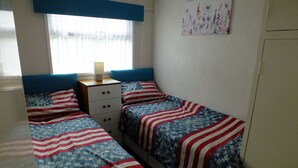 Une chambre à coucher avec deux lits, chacun recouvert d’une couverture aux motifs du drapeau américain, une commode et une fenêtre avec un rideau.