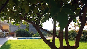A lush green lawn with a tree in the foreground, a pool in the background, and a building with outdoor seating. - Luxury Private Villa With Large Pool And Amazing Views  (Corinth)