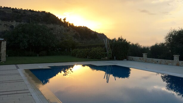 A rectangular swimming pool with a clear reflection of the sunset, surrounded by a paved deck and stone walls. - Luxury Private Villa With Large Pool And Amazing Views  (Corinth)
