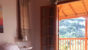 A bedroom with a bed, a nightstand, a lamp, a window with curtains, and a view of a balcony with a railing. - Luxury Private Villa With Large Pool And Amazing Views  (Corinth)