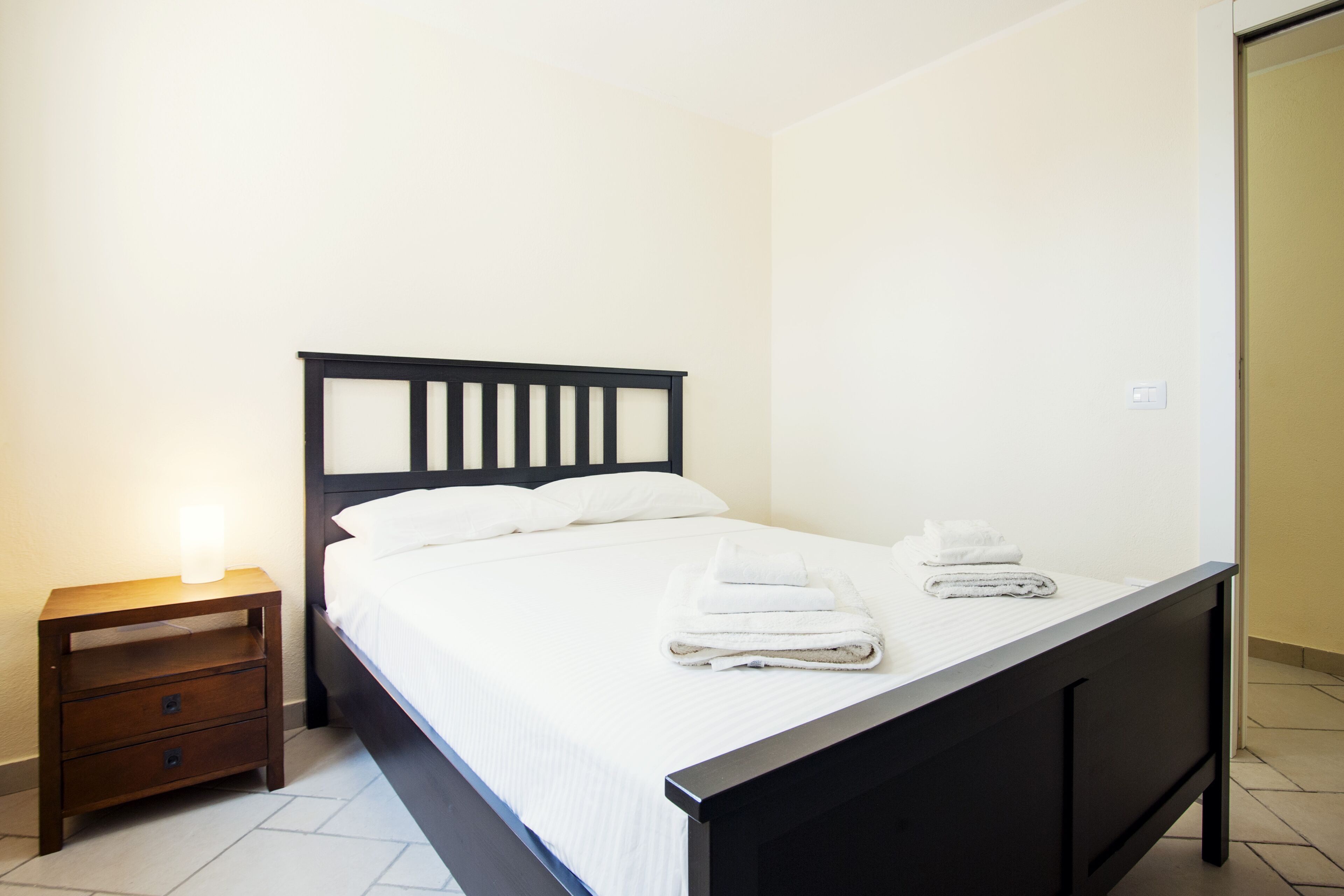Una cama bien tendida con sábanas blancas y dos toallas dobladas, una mesita de noche de madera con una lámpara, y un cabecero de madera oscura.