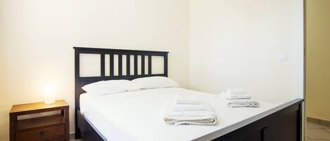 Una cama bien tendida con sábanas blancas y dos toallas dobladas, una mesita de noche de madera con una lámpara, y un cabecero de madera oscura.