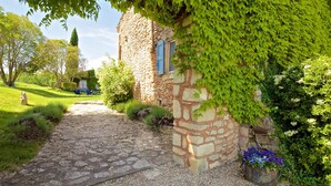 A stone building with ivy-covered walls, a gravel path, and a flower pot with purple flowers. - Beautifully renovated Artist’s farmhouse in Provence, large heated pool & sauna (Roussillon)