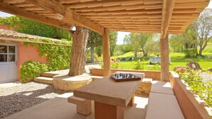 A covered outdoor area with a stone table and benches, surrounded by greenery and a view of a garden. - Beautifully renovated Artist’s farmhouse in Provence, large heated pool & sauna (Roussillon)