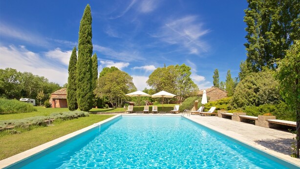 A rectangular swimming pool with clear blue water, surrounded by lounge chairs and umbrellas, set in a lush garden with tall trees and a clear sky. - Beautifully renovated Artist’s farmhouse in Provence, large heated pool & sauna (Roussillon)