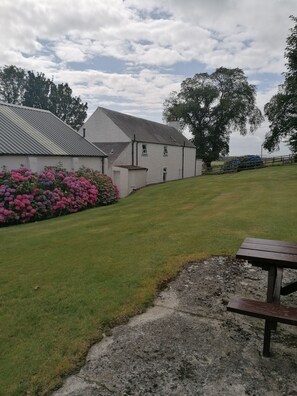 A white building with a sloped roof, surrounded by a grassy area and pink flowers. - Dairy Cottage East Challoch Farm (Stranraer)