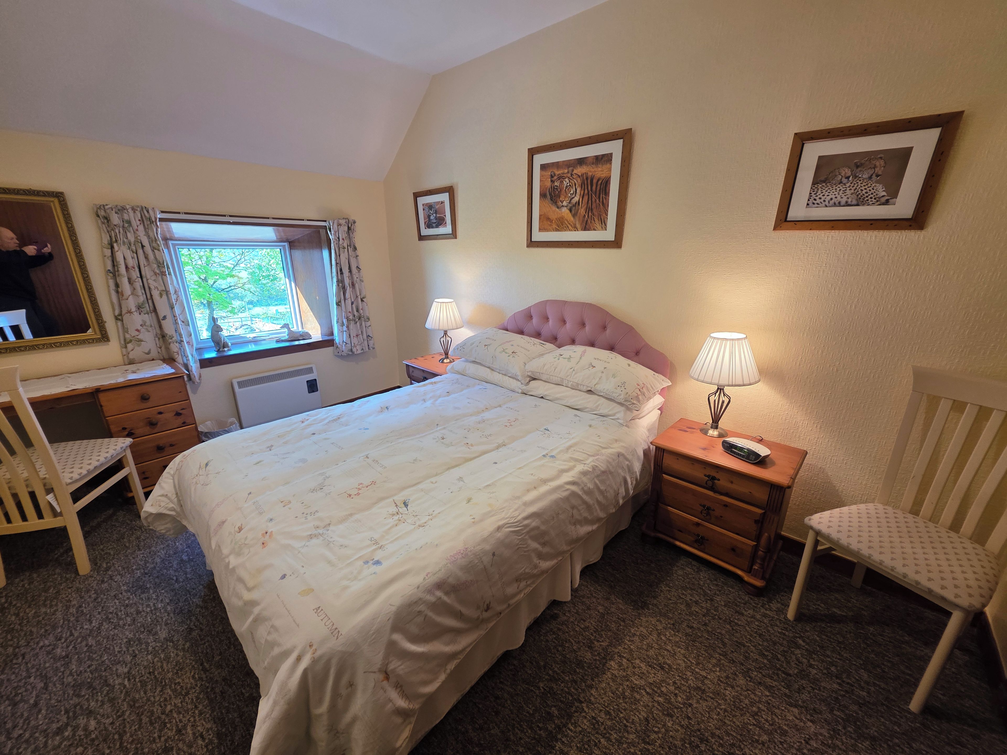 A bedroom with a bed, two bedside tables, a chair, a dresser, a window with curtains, and wall-mounted pictures.