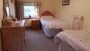A bedroom with a wooden dresser, a bedside table with a lamp, a window with curtains, a bed with a floral bedspread, and a chair. - Dairy Cottage East Challoch Farm (Stranraer)