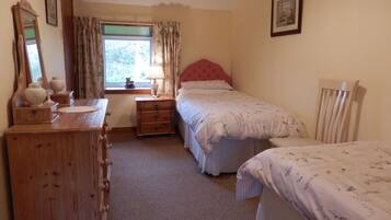 A bedroom with a wooden dresser, a bedside table with a lamp, a window with curtains, a bed with a floral bedspread, and a chair.
