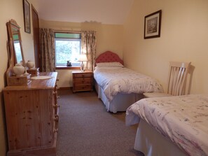 A bedroom with a wooden dresser, a bedside table with a lamp, a window with curtains, a bed with a floral bedspread, and a chair. - Dairy Cottage East Challoch Farm (Stranraer)