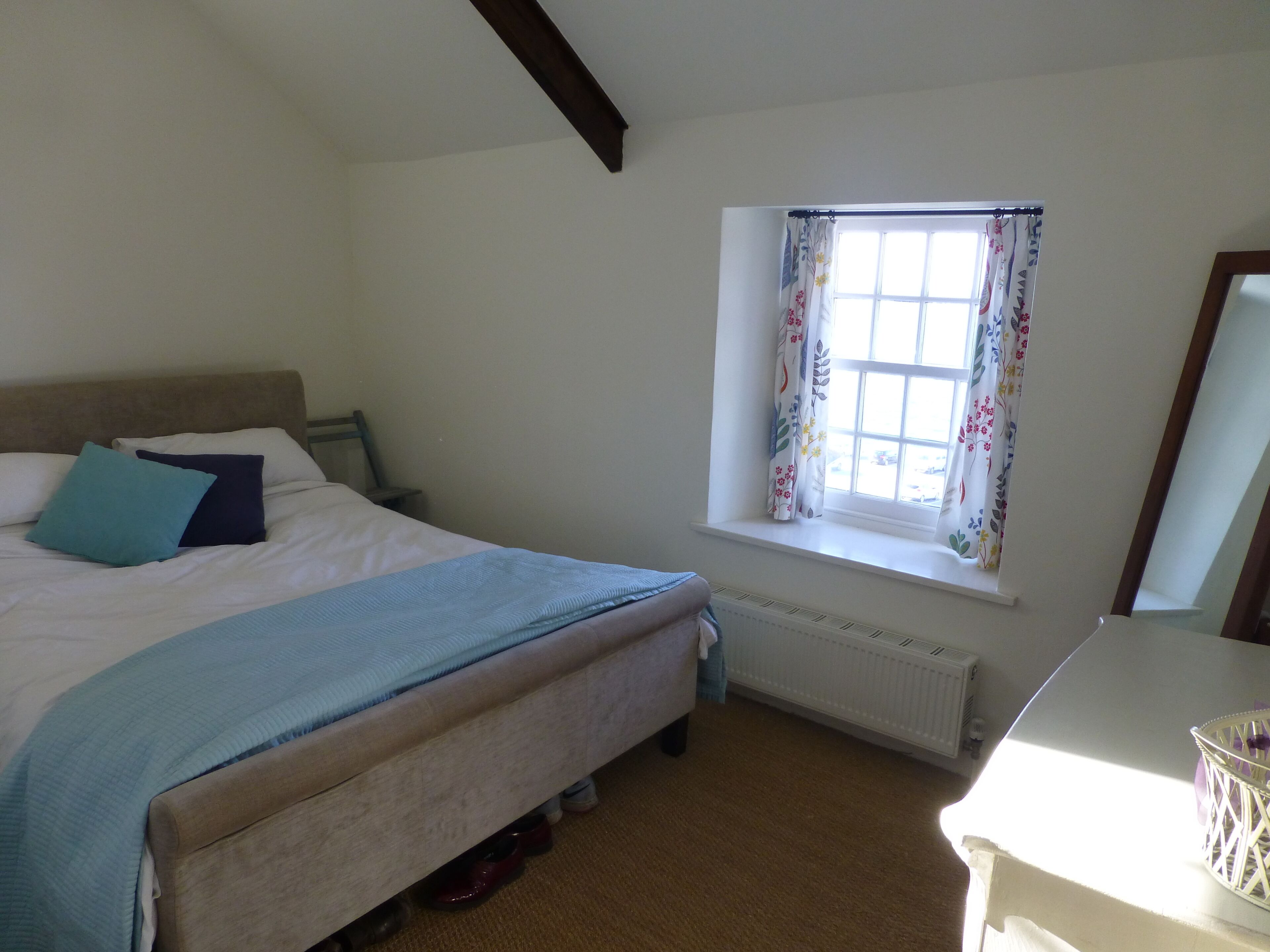 A bedroom with a bed, a window with floral curtains, a radiator, and a dresser.