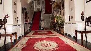Un amplio pasillo con alfombra roja, sillas de madera, una escalera y una cabeza de venado montada.