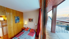 Ein Zimmer mit Holzwänden, einem Bett mit gemustertem Überwurf, einem Gemälde, einem gerahmten Bild und Blick auf eine verschneite Landschaft vom Balkon aus.