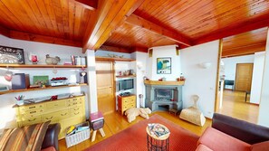 Ein gemütliches Wohnzimmer mit Holzbalken, Kamin, Fernseher und Bücherregal.