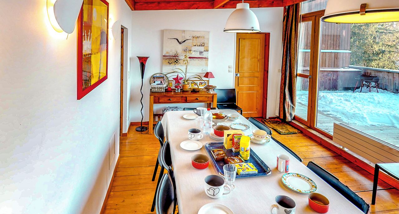 A dining area with a long table set for four, a wooden floor, and a large window with a view of a snowy landscape.