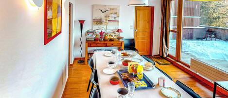 A dining area with a long table set for four, a wooden floor, and a large window with a view of a snowy landscape.