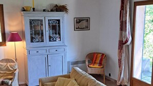 A cozy living room with a beige sofa, a white cabinet with glass doors, a wicker chair, and a view of the outdoors through a window with curtains. - Lovely Family Home With Pool, Complete Privacy And Views Over The Lubero  (Forcalquier)