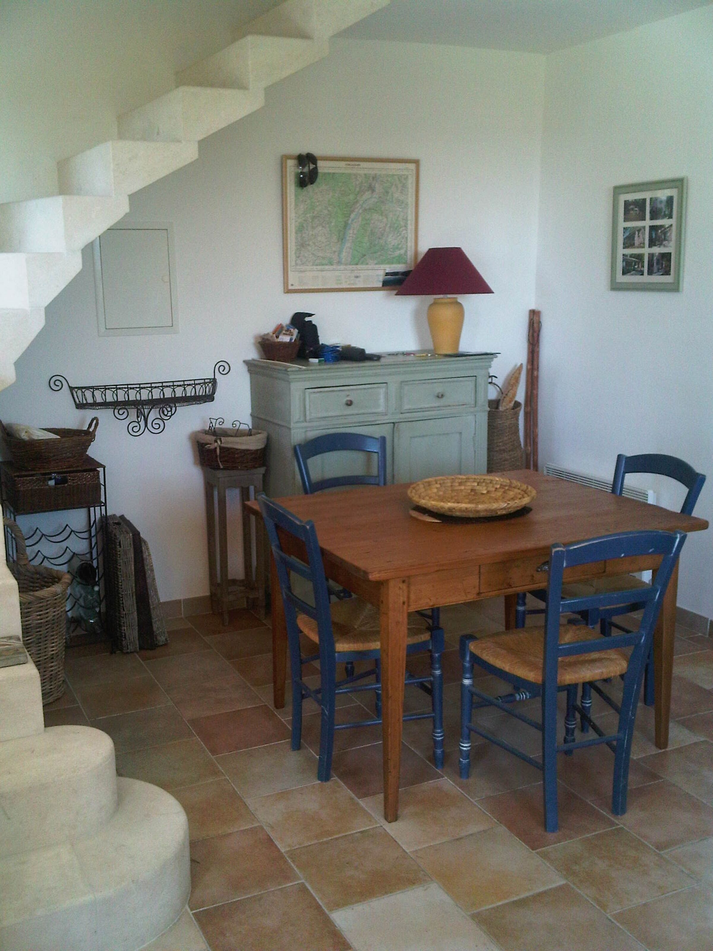 Un comedor con mesa de madera y sillas azules, una escalera, un mapa en la pared y una lámpara sobre un aparador.