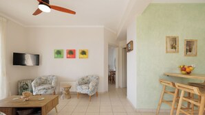 A living room with a TV, two patterned chairs, a wooden coffee table, and a dining area with a fruit bowl.
