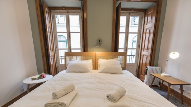 Habitación de hotel con una cama grande, dos almohadas, una mesita de noche con lámpara y vistas a través de una ventana con marcos de madera.