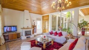 A living room with a fireplace, a sofa set, a television, and a large window. - Villa Lira | 6 bedrooms | Spacious | Quinta do Lago (Quinta do Lago)